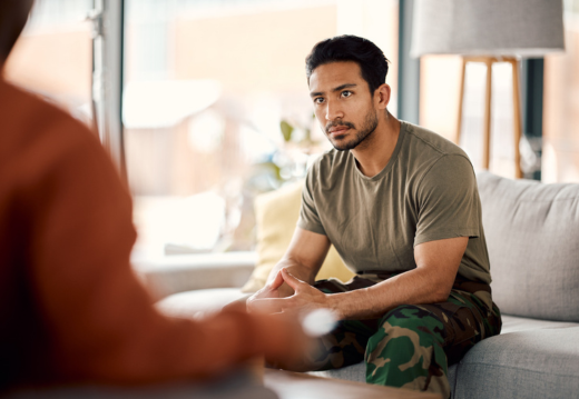 Young male veteran in camouflage pants and a t-shirt sits on a couch in a therapy session, attentively listening and engaged in a mental health consultation.
